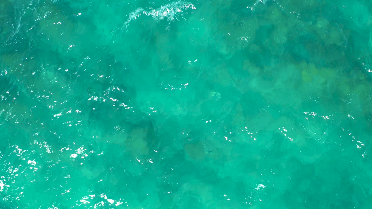 Overhead view of clear turquoise water in East Cape, Baja California Sur, with a rocky seabed visible under small ripples and no visible people.