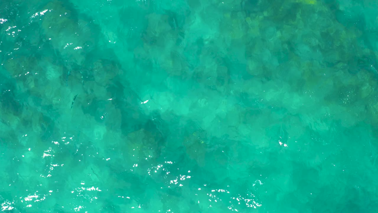 Turquoise ocean water in East Cape, Baja California Sur, fills the frame with scattered white reflections and mottled rocky seabed visible below, with no visible people.