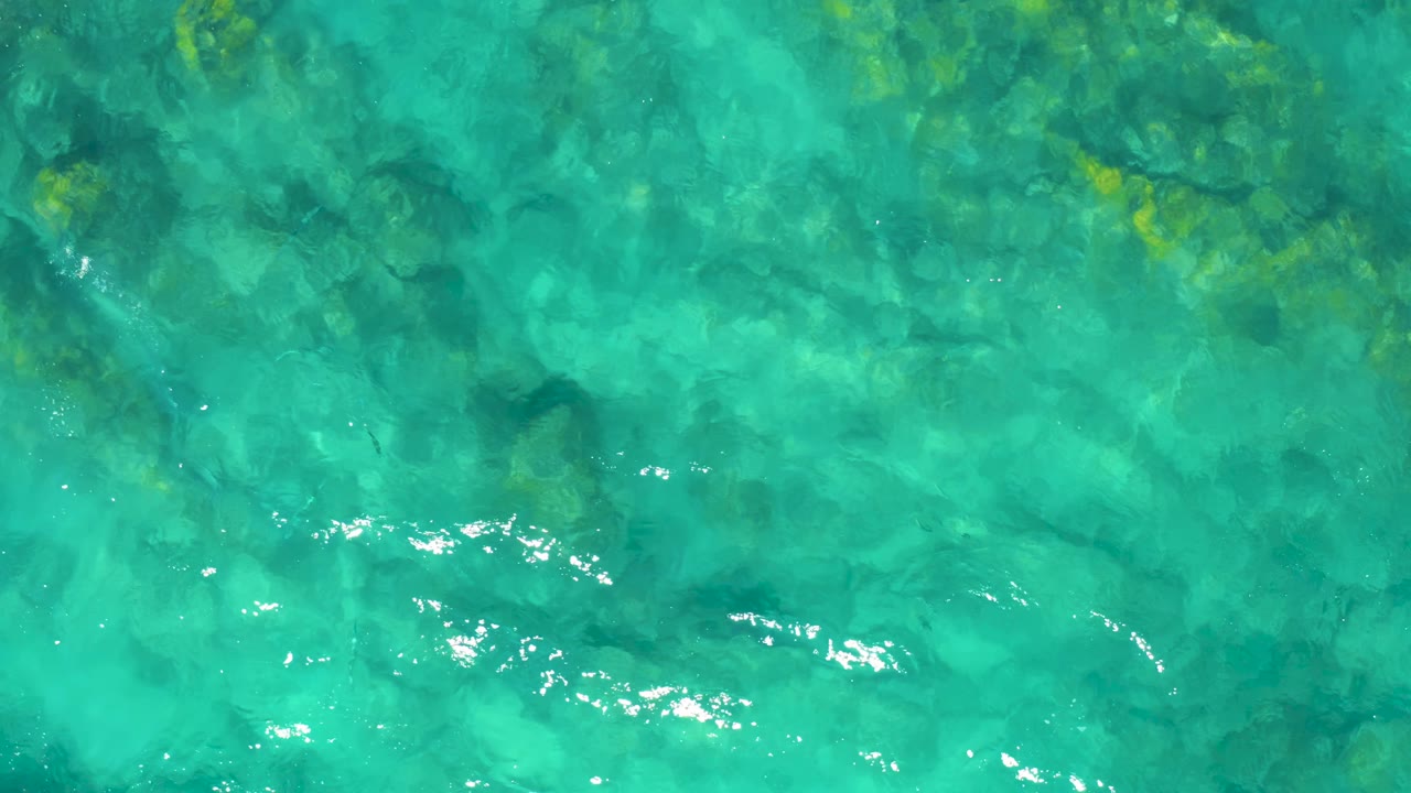 Aerial frame of clear turquoise water with mottled submerged rocks and small white light reflections, with no visible people.
