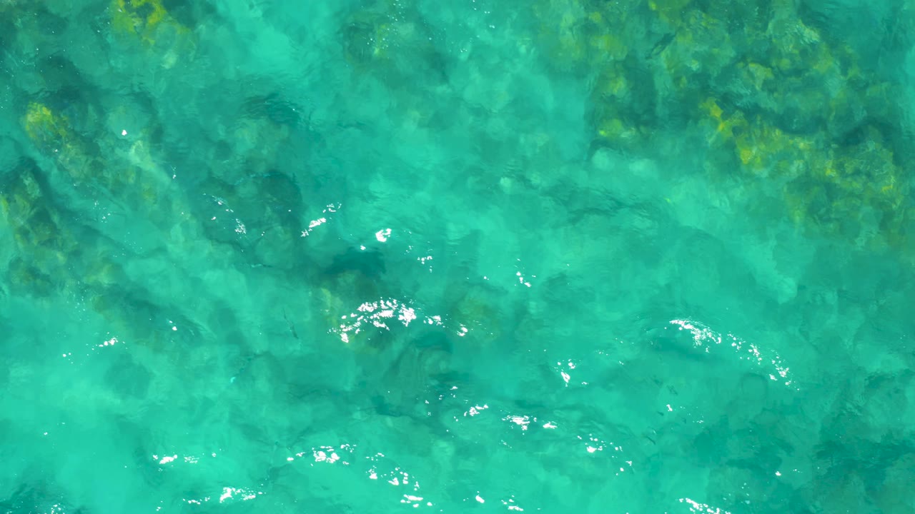 Top-down view of shallow turquoise ocean where darker rocks are visible below the surface and scattered sun glints cross the water, with no visible people.