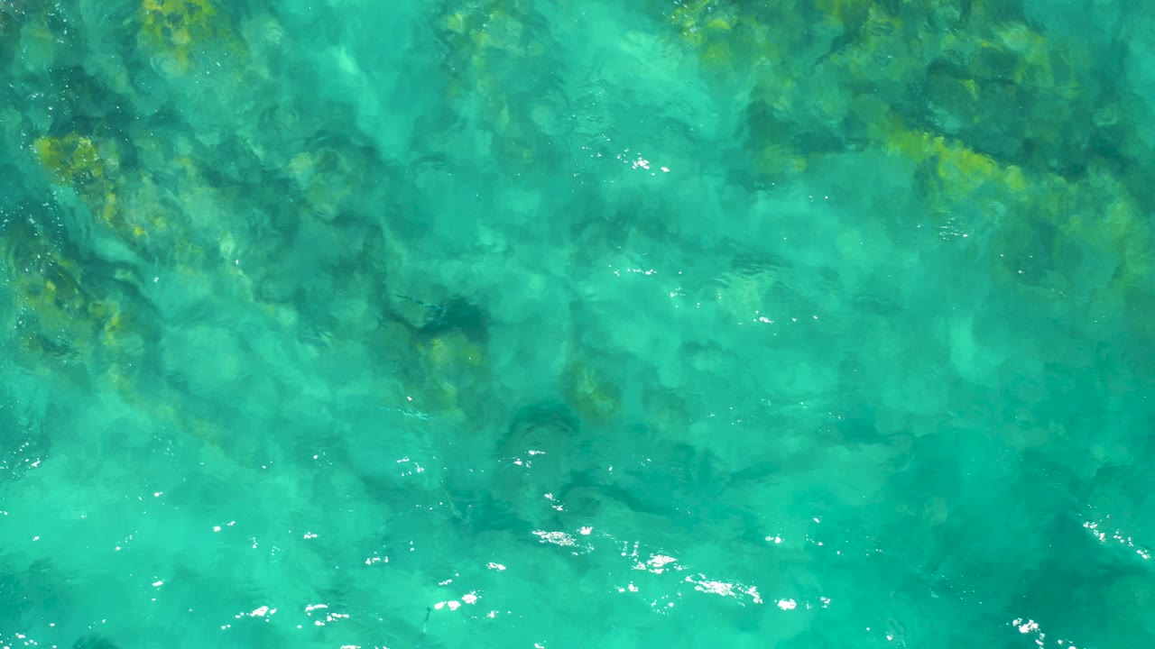 Aerial ocean frame in East Cape, Baja California Sur showing turquoise water, submerged rocks, and a brighter patch of white foam forming near the center, with no visible people.