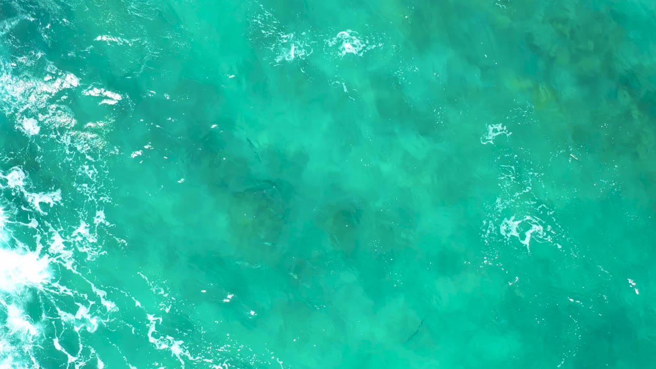 Aerial frame over clear turquoise water in East Cape, Baja California Sur, where white foam gathers near the lower left and darker underwater shapes show through, with no visible people.