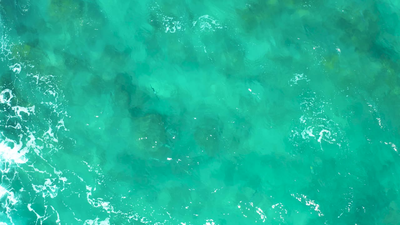Straight-down East Cape ocean view with transparent turquoise water, scattered white foam lines on the left and upper right, visible seabed color variation, and no visible people.