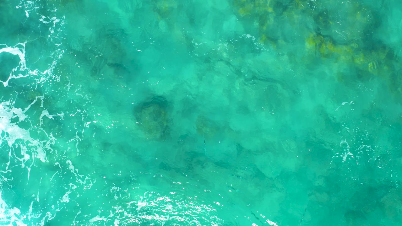 Top-down view of turquoise ocean water in East Cape, Baja California Sur, with white foam entering from the left edge, darker underwater rocks below the surface, and no visible people.