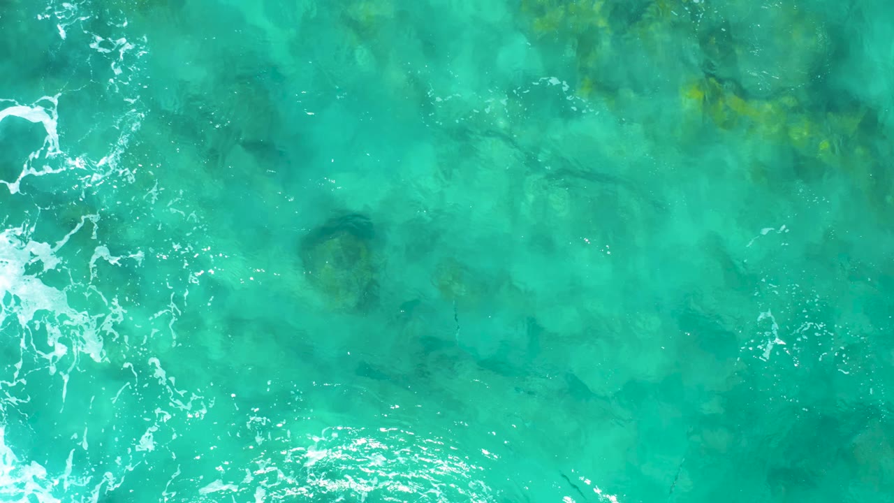 Clear turquoise sea surface with submerged rocks and small foam streaks.