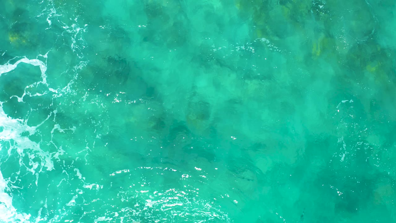 Aerial view over East Cape turquoise water as a brighter white foam line curls along the left side above visible seabed and underwater rock forms, with no visible people.