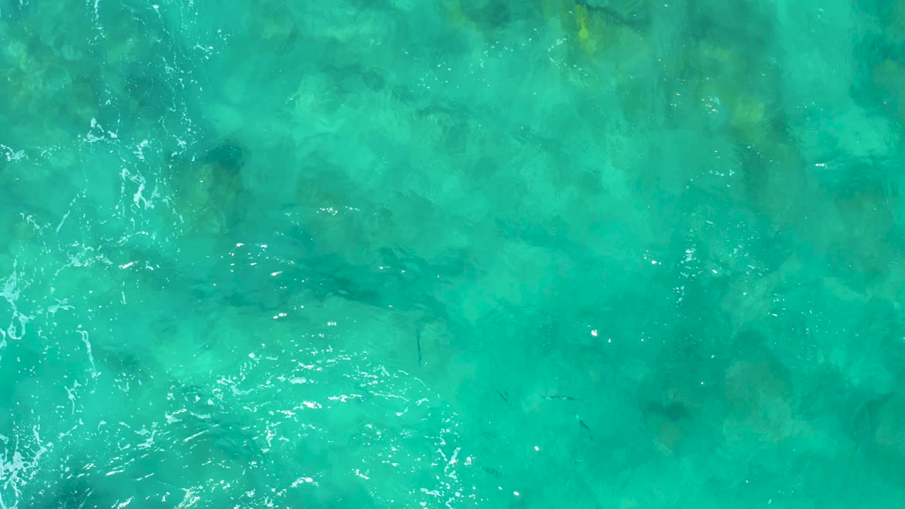 Aerial view of green-blue ocean water in East Cape BCS Mexico, with mottled seabed shapes beneath the surface and a cluster of bright ripples near the lower edge, with no visible people.