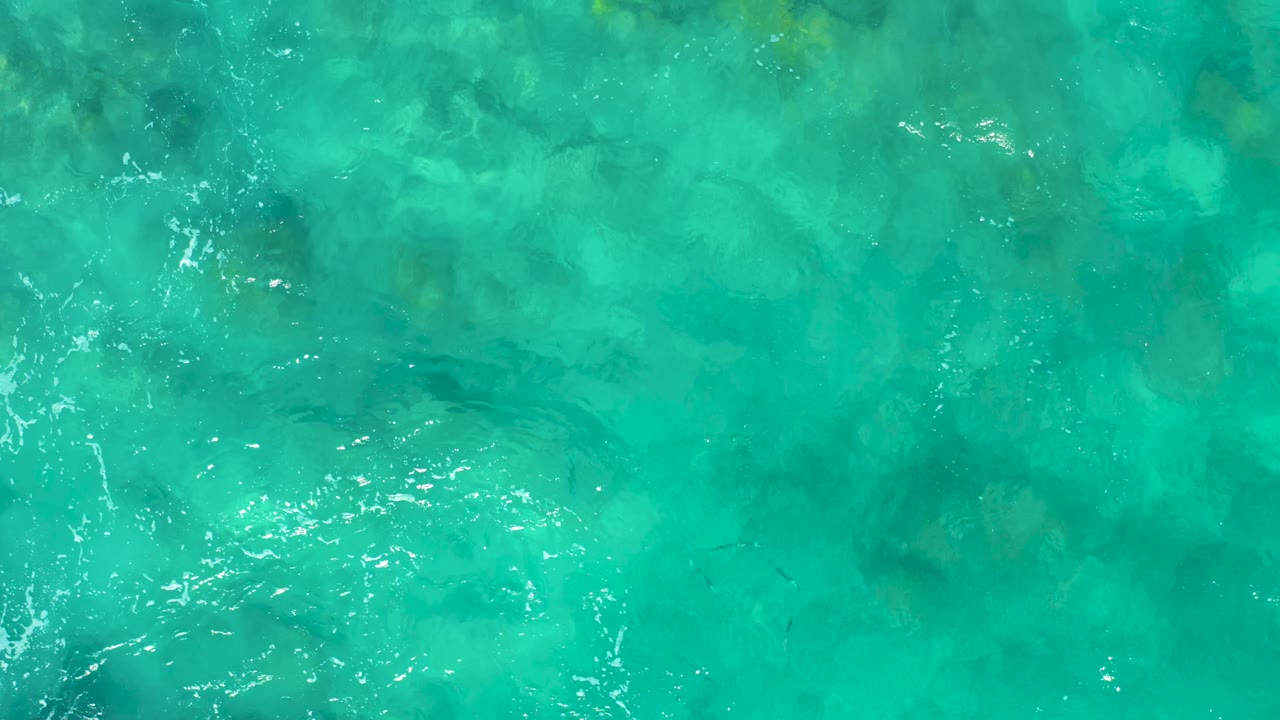 Top-down aqua ocean view in East Cape BCS Mexico, showing darker underwater patches, pale sandy seabed color, and small white highlights from surface ripples, with no visible people.