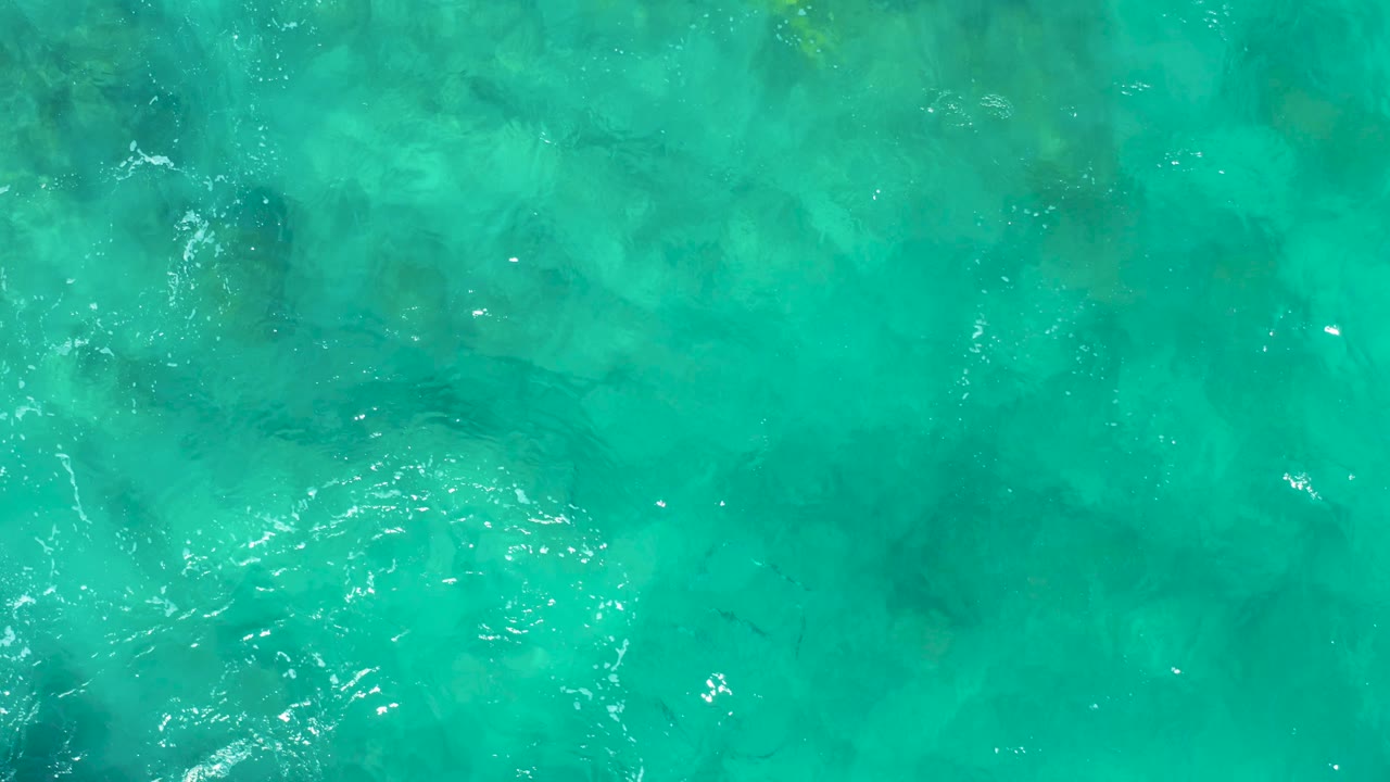 Overhead frame of shallow turquoise water in East Cape BCS Mexico, with blurred rock or seabed forms under the surface and thin ripple lines crossing the left side, with no visible people.