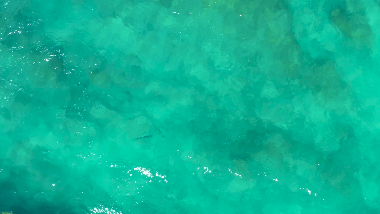 Aerial view of turquoise ocean water in East Cape, Baja California Sur, Mexico, with darker seabed patches visible beneath the rippled surface and no visible people.