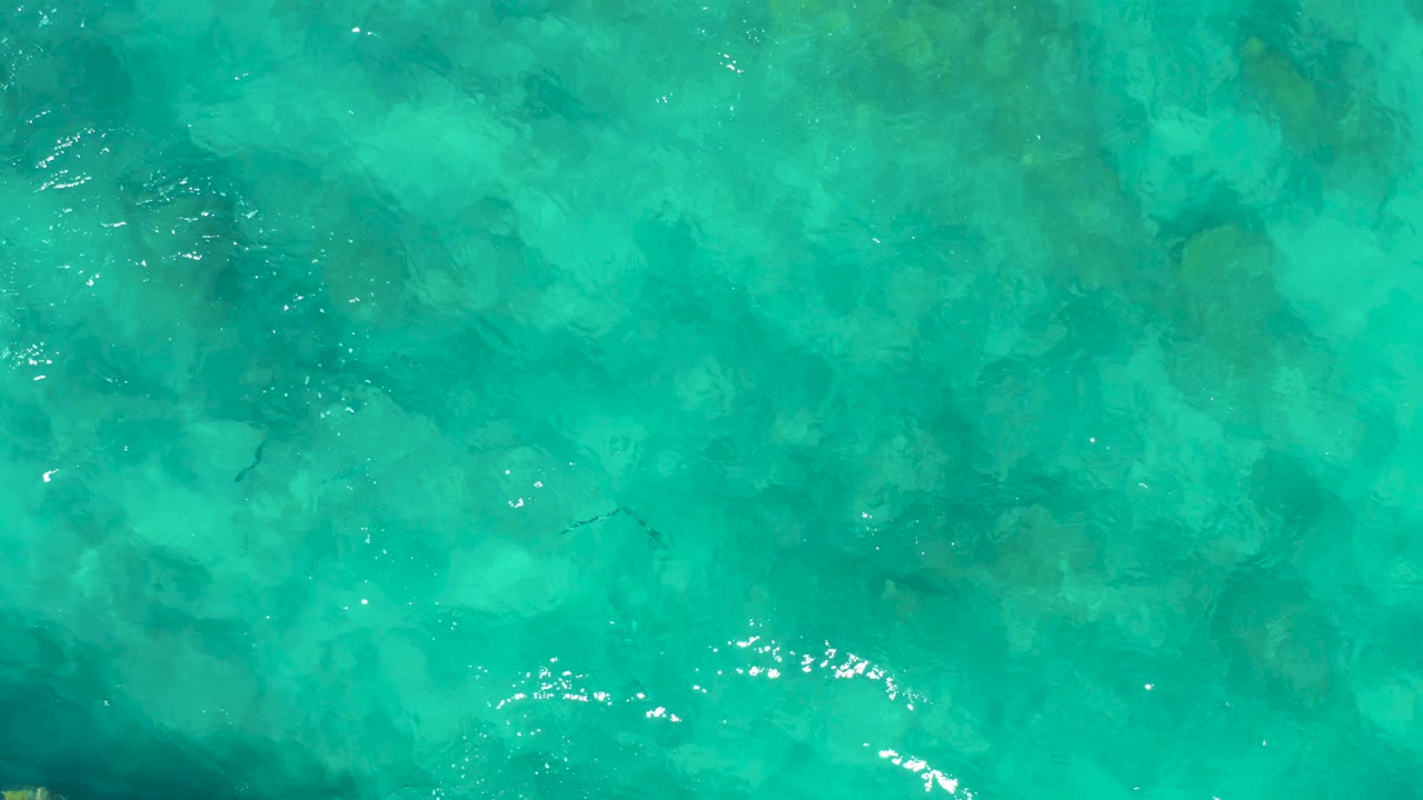 Shallow turquoise water over a mottled seabed in East Cape, Baja California Sur, Mexico, with faint small fish shapes and no visible people.