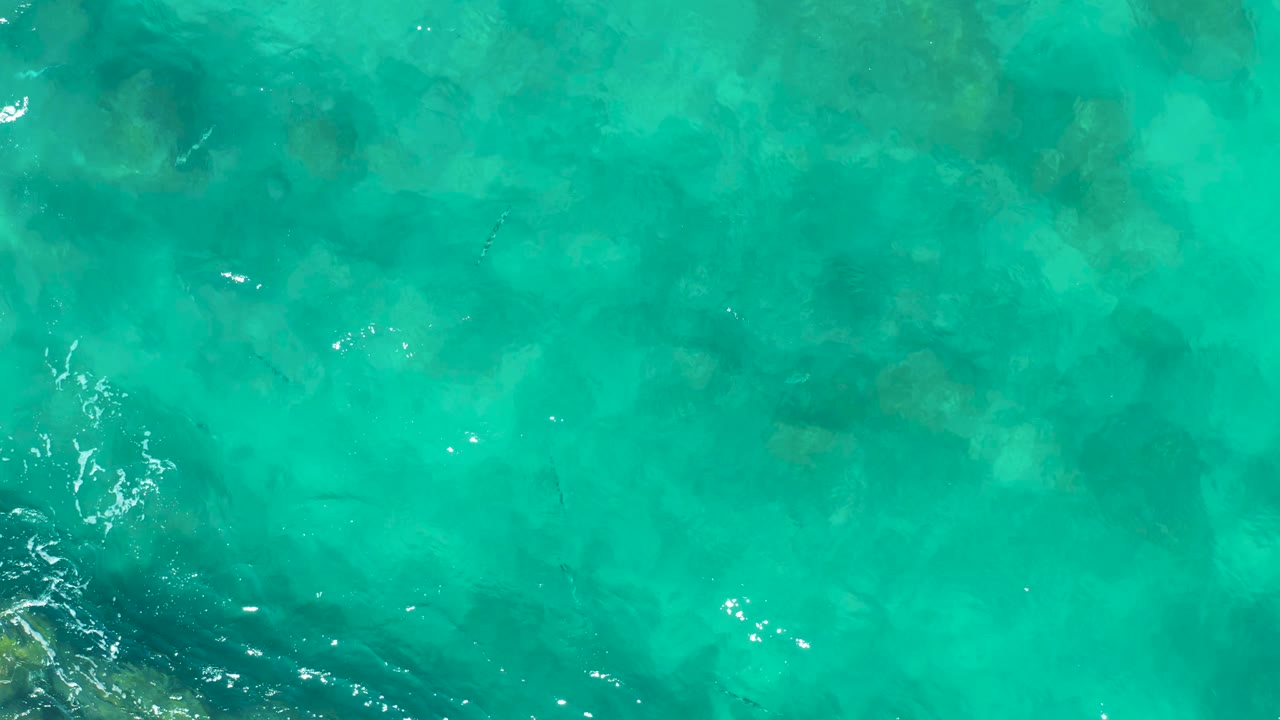 Aerial view of clear green-blue East Cape, Baja California Sur water, with underwater rock shapes visible through the surface and brighter ripple highlights clustered toward the lower left, no visible people.