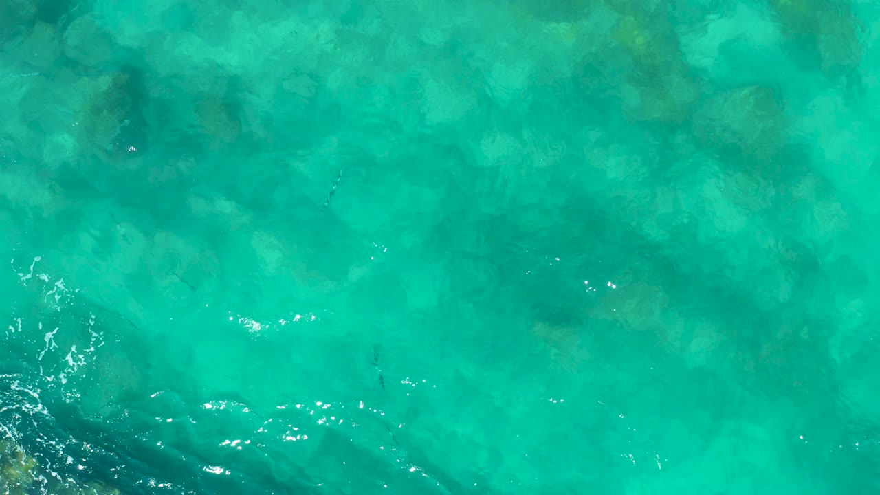 Top-down frame of turquoise East Cape, Baja California Sur ocean with darker underwater rock patches along the lower left and scattered white surface glints, no visible people.