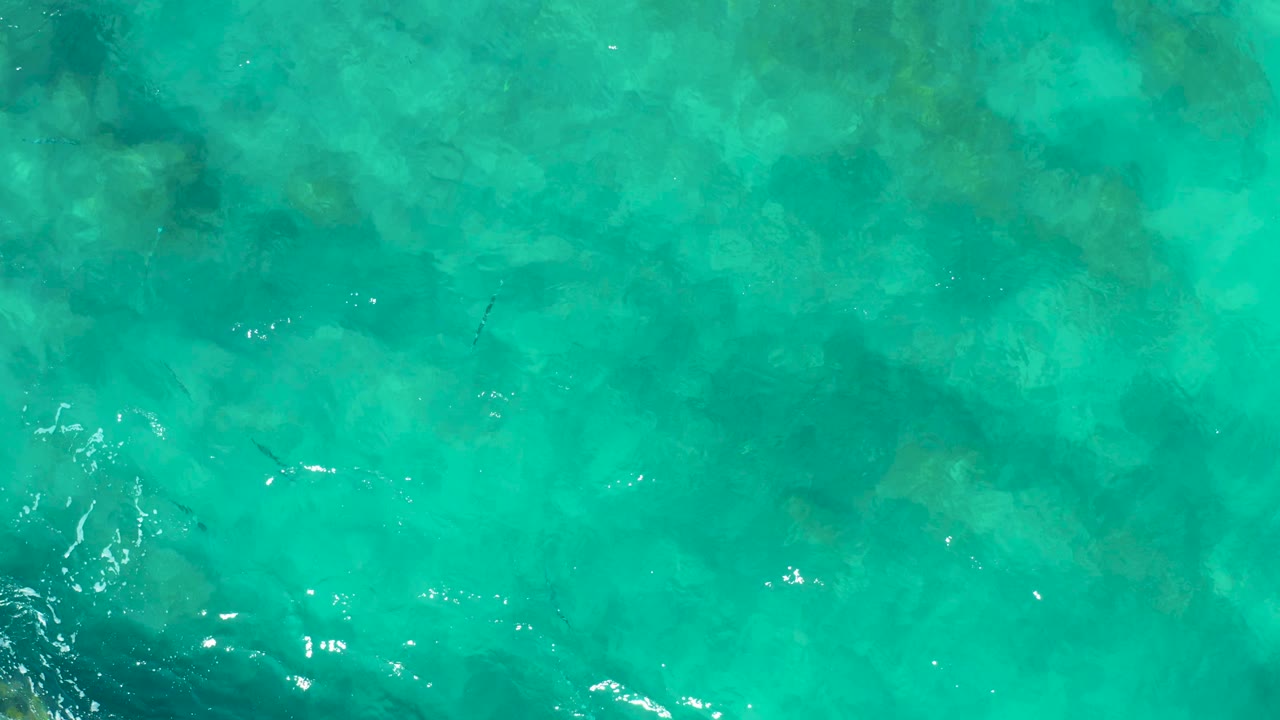 Aerial frame over shallow East Cape, Baja California Sur water, showing mottled submerged rocks across the center and small ripples near the lower edge, no visible people.