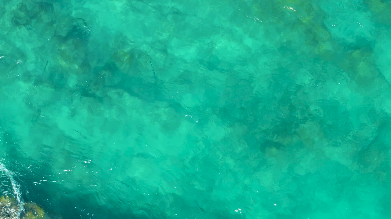Top-down view of turquoise East Cape BCS Mexico ocean water with submerged rocks, faint surface ripples, and no visible people.