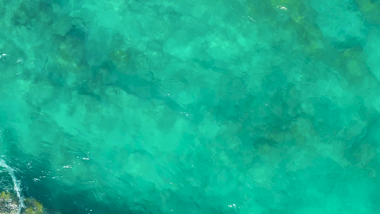 Overhead frame of East Cape BCS Mexico turquoise water where darker submerged rock shapes sit beneath gentle ripples, with no visible people.