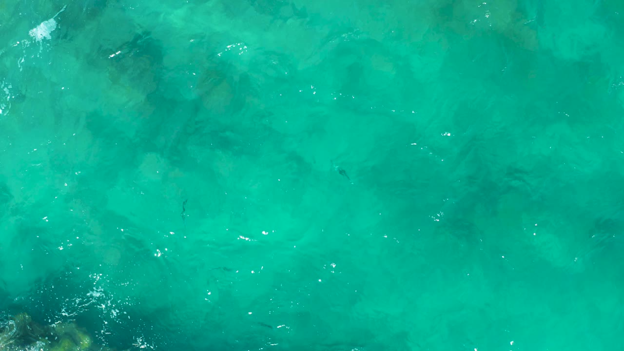 Aerial frame over East Cape, Baja California Sur, with turquoise water, darker submerged reef areas, a few white foam marks near the top, and no visible people.