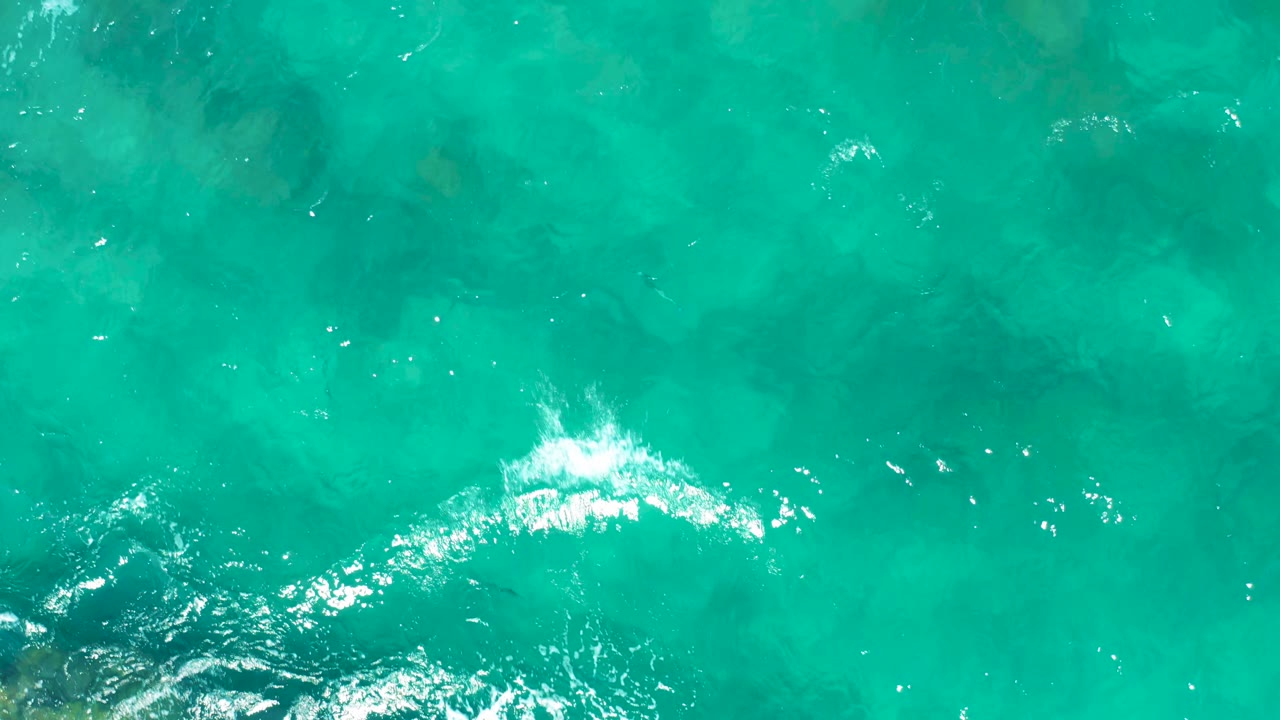 Overhead view of shallow East Cape water with a brighter band of white wave foam crossing turquoise surface above visible rocks, with no visible people.