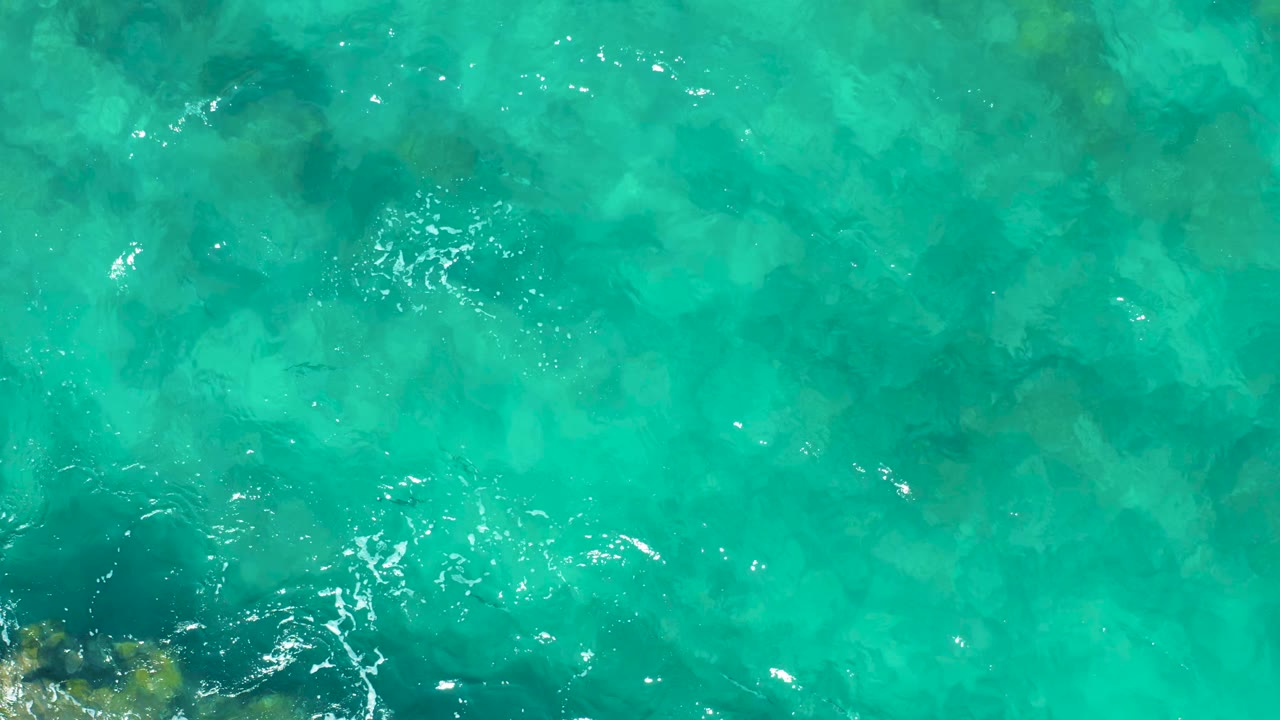 Aerial frame over East Cape turquoise water with submerged rocky texture, faint green-blue seabed variation, and scattered white ripples, no visible people.