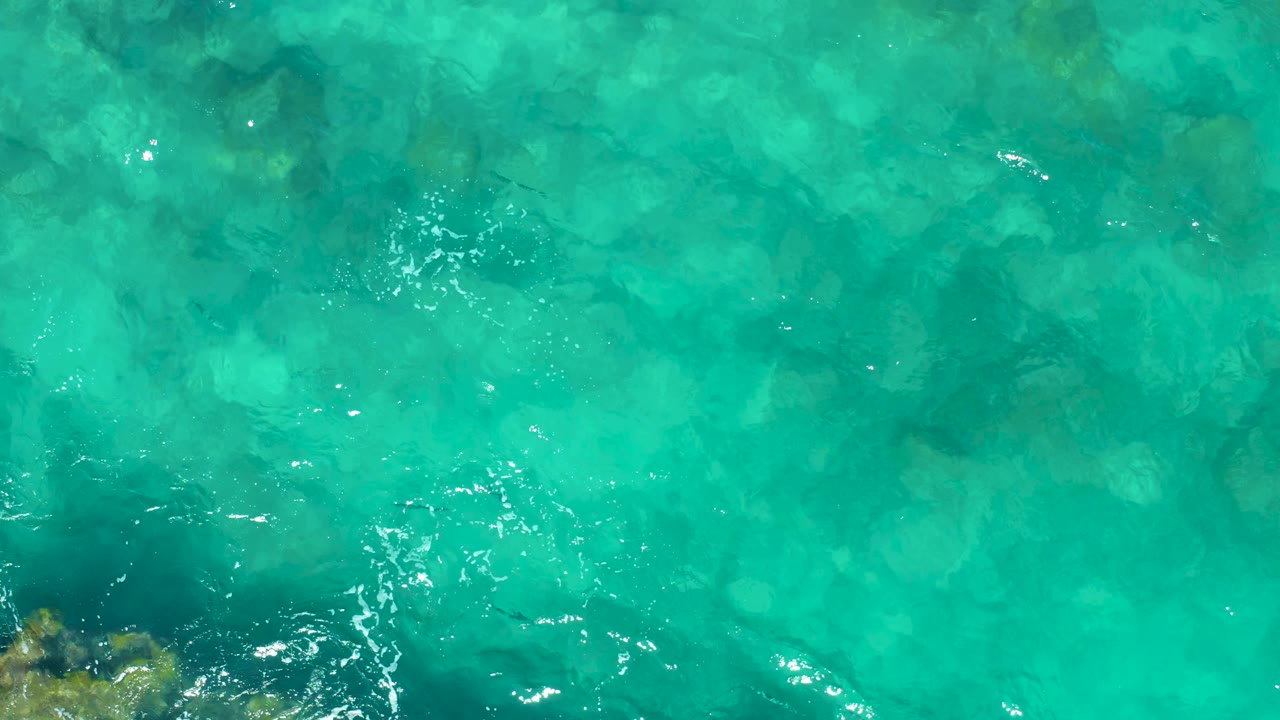 Overhead East Cape ocean frame showing a darker underwater patch near the lower center, bright ripple marks, and transparent turquoise water, no visible people.