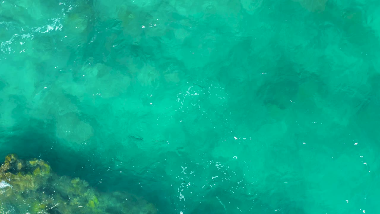 Aerial frame of clear green-blue ocean water in East Cape, Baja California Sur, with a rocky outcrop partly visible along the lower left and no visible people.
