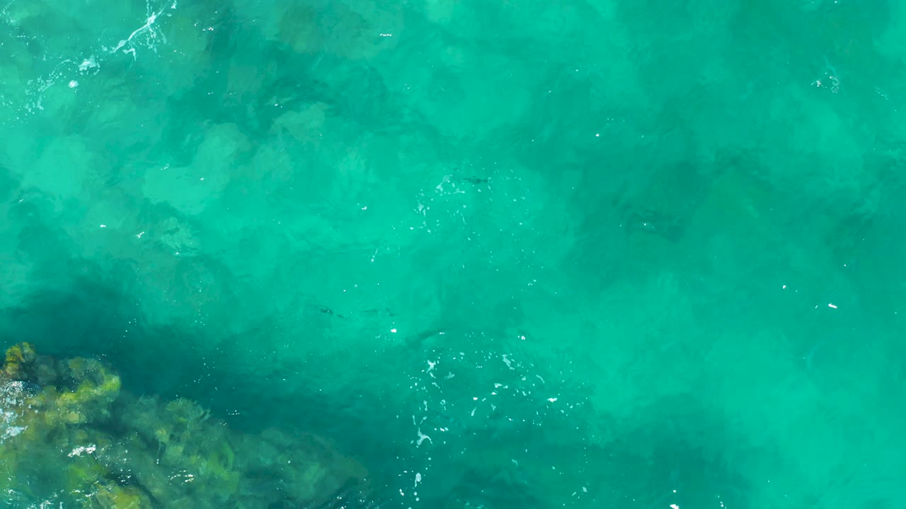 Top-down view of turquoise ocean in East Cape, Baja California Sur, with underwater rocks entering from the bottom edge and no visible people.