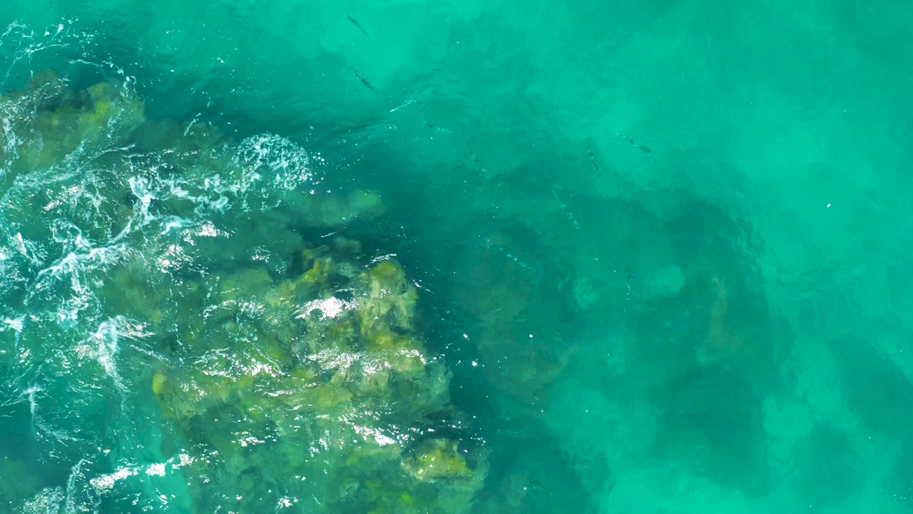 East Cape BCS Mexico shallows seen from above, with a larger rock mass low in frame and white water skimming the left edge, no visible people.