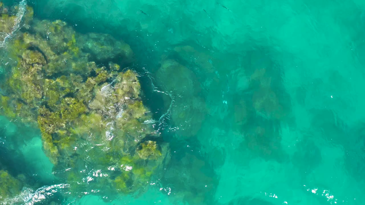 Clear green-blue water in East Cape, Baja California Sur, Mexico, shows the main underwater rock mass along the left edge with faint darker formations to the right and no visible people.