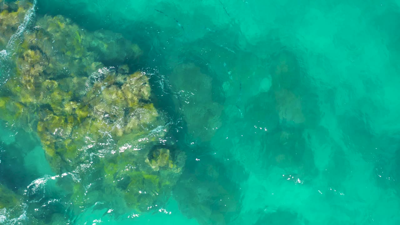 Aerial top-down frame from East Cape, Baja California Sur, Mexico, with rocky underwater formations near the upper left and bright turquoise water stretching across the right, with no visible people.