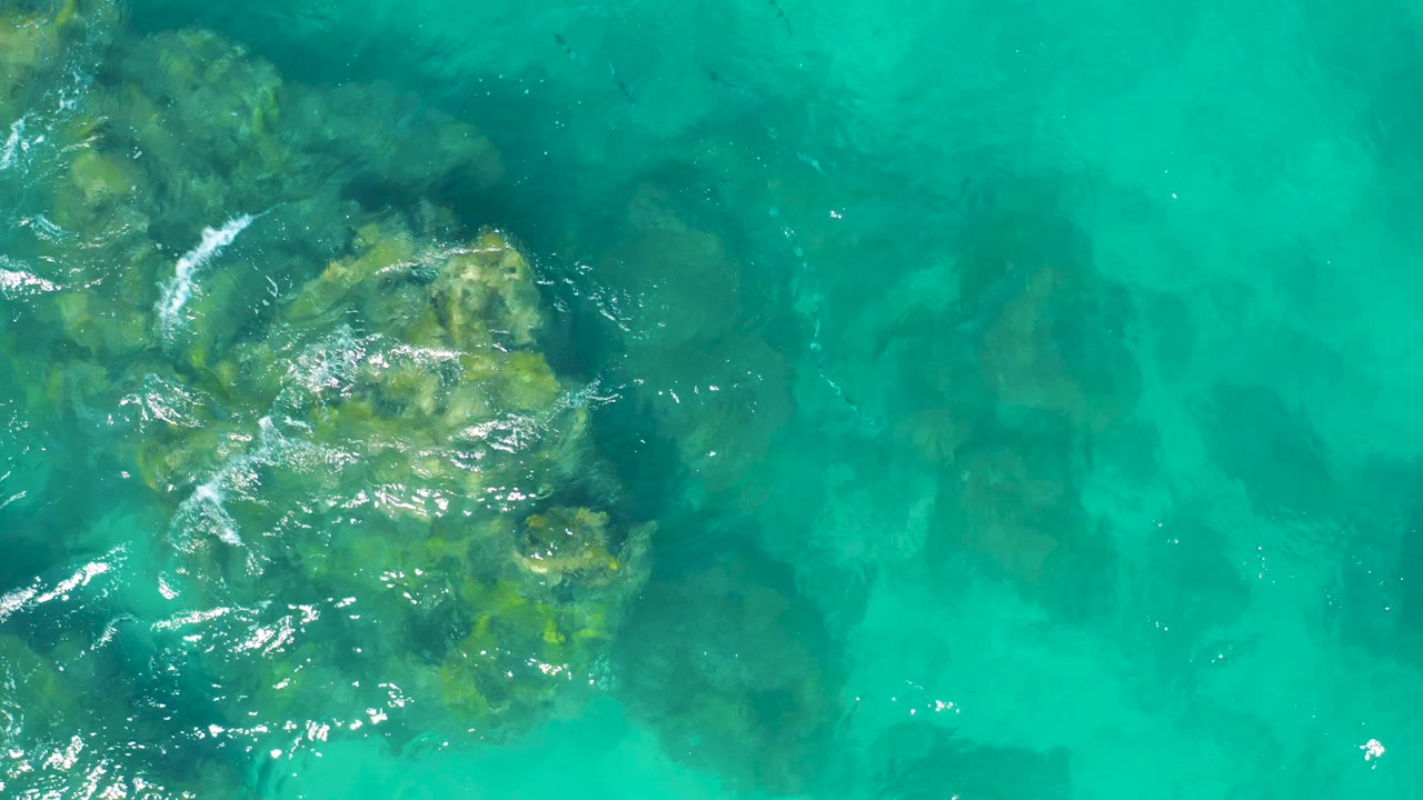 Top-down East Cape, Baja California Sur, Mexico frame showing a large submerged rock mass in the lower left, darker water above it, and open turquoise water to the right with no visible people.