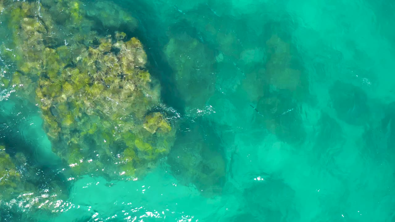 Aerial frame over East Cape Baja California Sur water with the main submerged rock still left of center, brighter glints along the lower edge, and no visible people.