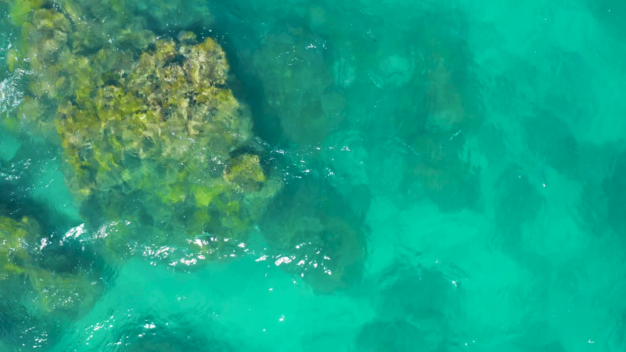 East Cape Baja California Sur aerial view with an algae-covered rock mass along the left side, transparent water over darker seabed patches, and no visible people.