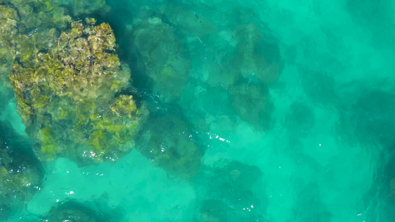 Aerial frame over East Cape, Baja California Sur water with the rock outcrop still near the left side, dark underwater shapes across the center, and no visible people.