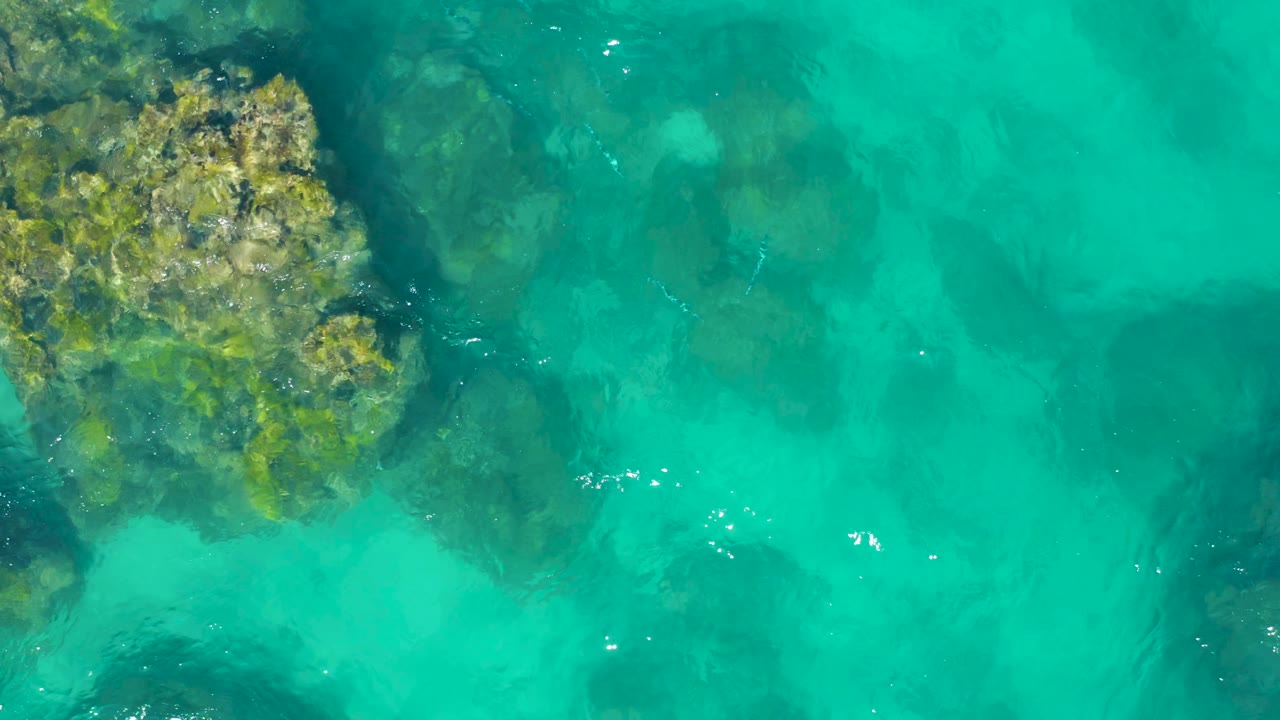 Top-down view of East Cape, Baja California Sur clear water, with a rough rock edge at upper left, muted underwater rocks through the center, and no visible people.