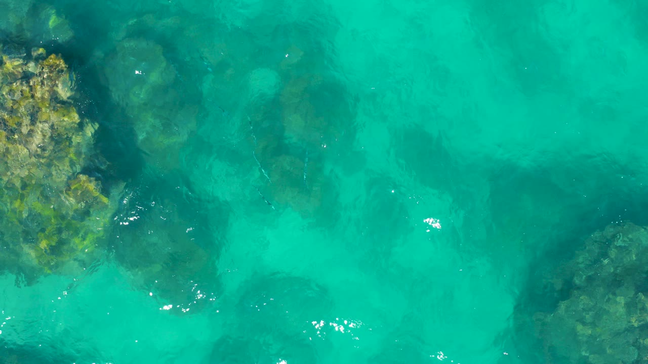 Overhead view of clear turquoise water over dark submerged rocks in East Cape BCS Mexico, with small surface highlights and no visible people.