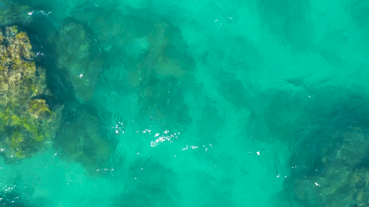 Top-down view of shallow turquoise water in East Cape BCS Mexico, with darker seabed patches and scattered bright ripple reflections, no visible people.