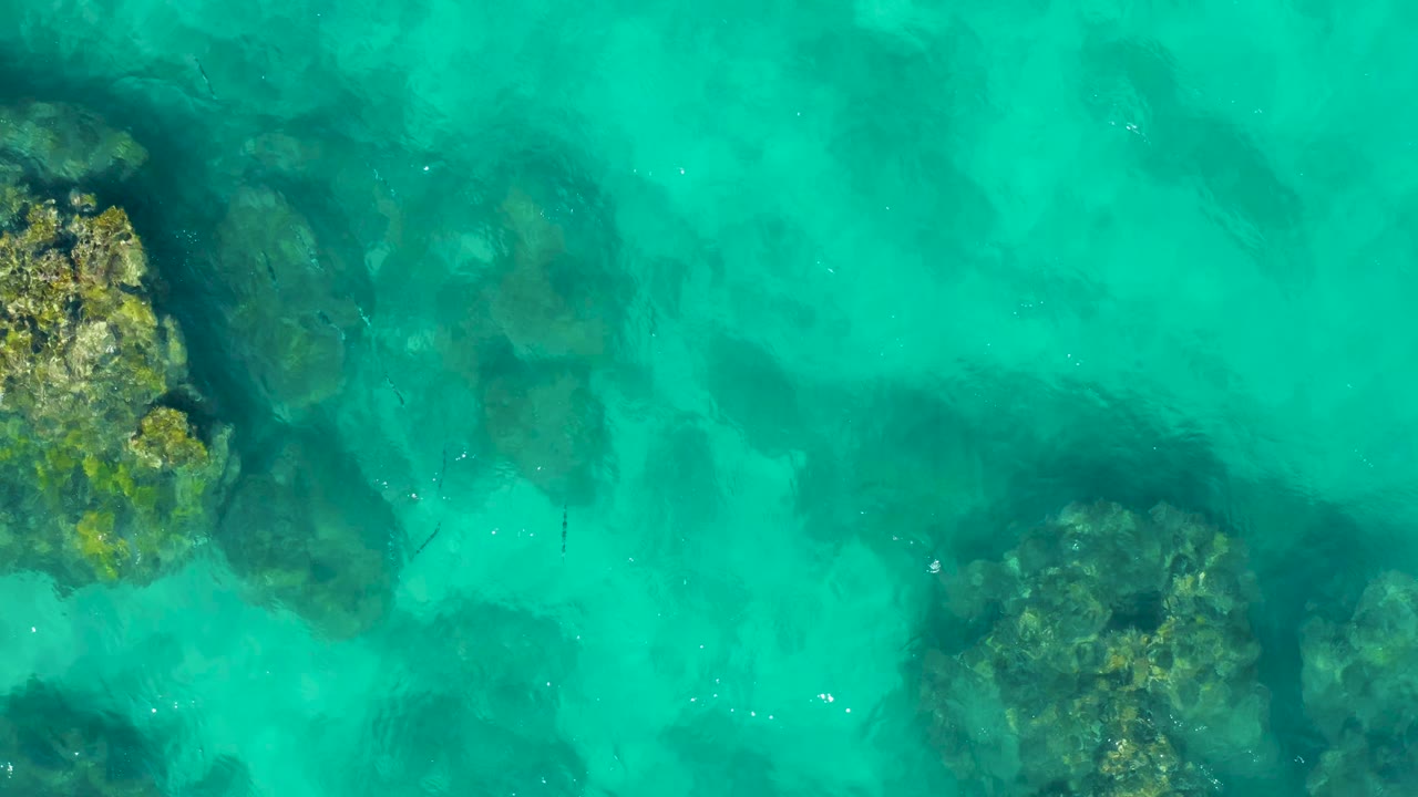 East Cape, Baja California Sur aerial frame with turquoise water, a rough rock formation along the left edge, darker underwater patches near center, and no visible people.