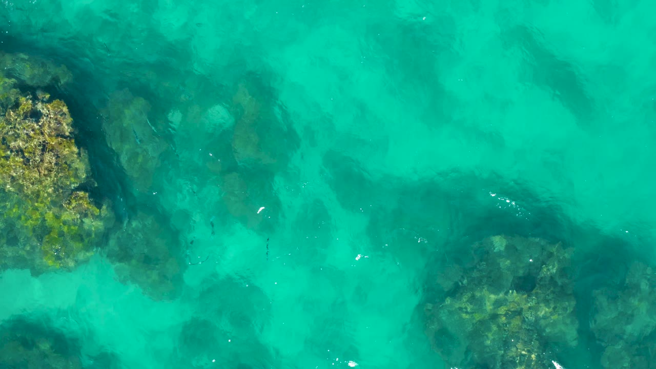Overhead East Cape, Baja California Sur water detail with a rock cluster at upper left, another submerged rock area at lower right, scattered white surface reflections, and no visible people.