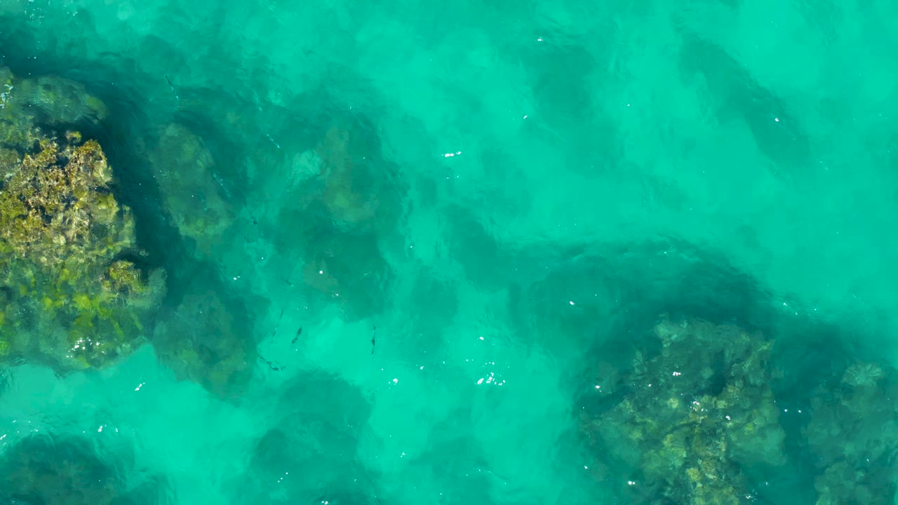 East Cape, Baja California Sur shallows seen from above, with a rocky patch at left, darker submerged forms across the middle, soft ripples, and no visible people.