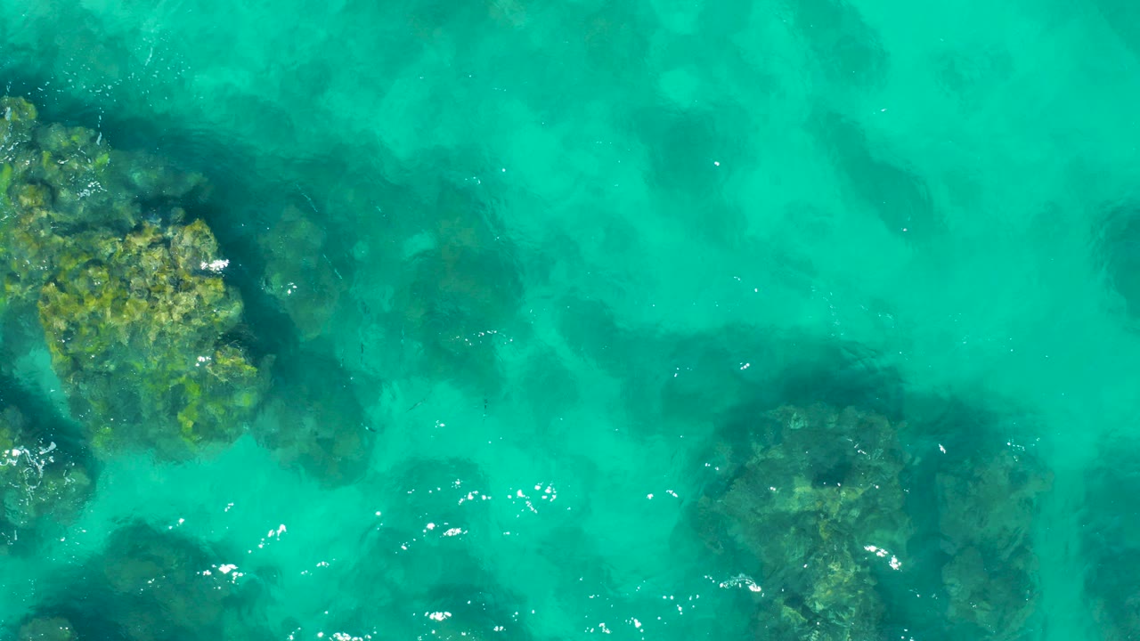 Overhead view in East Cape, Baja California Sur, showing clear turquoise water with dark submerged rocks and small white ripples, no visible people.