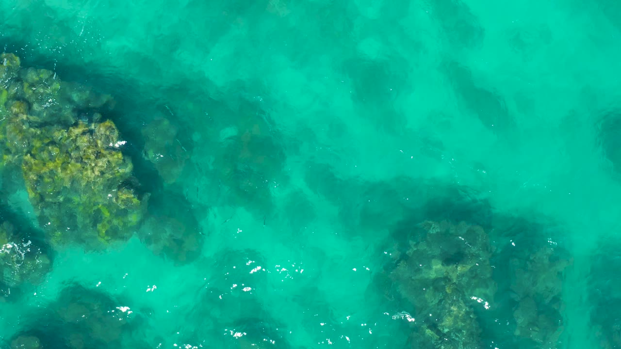 Overhead East Cape frame with a large rocky patch at upper left, darker seabed forms below the surface, and light foam marks across turquoise water, no visible people.