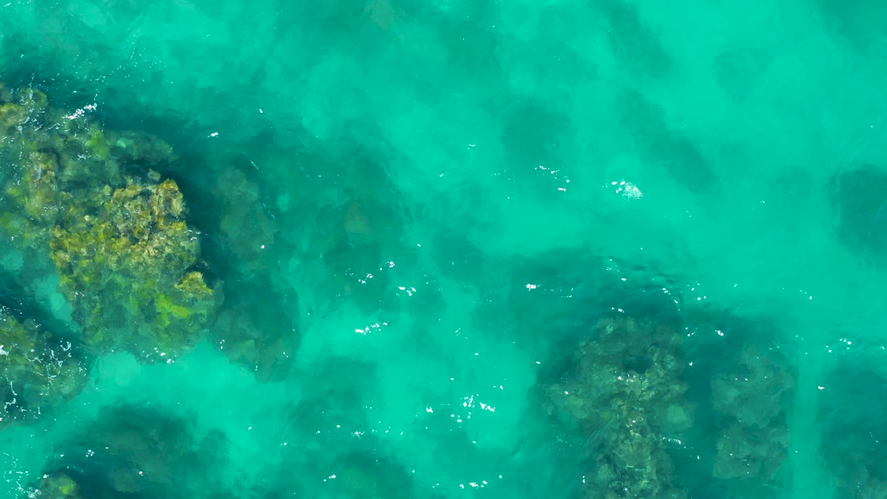 Overhead view of East Cape water with a greenish rock cluster near the upper left and a rounded submerged rock area at right under brighter turquoise current, no visible people.