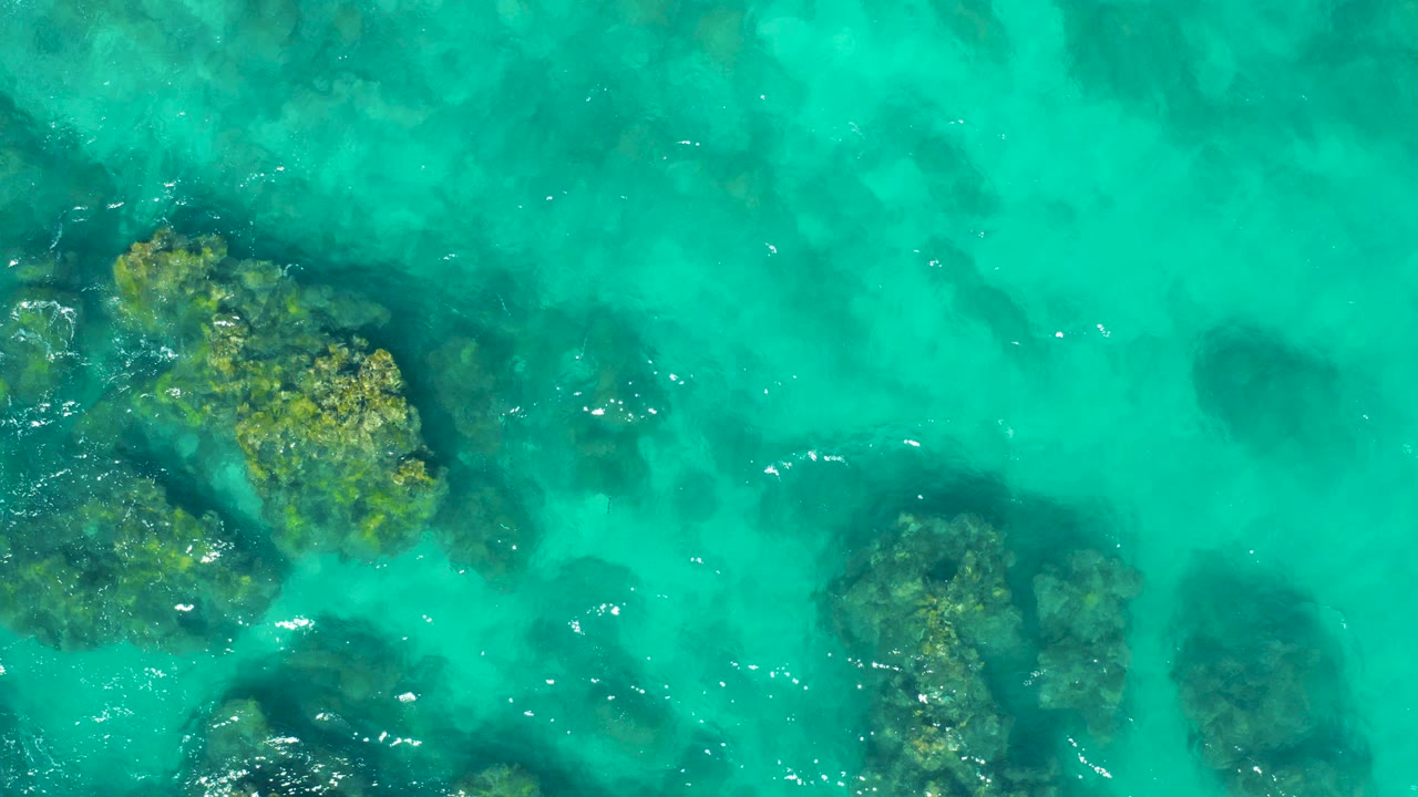Overhead view of clear turquoise water in East Cape, Baja California Sur, with dark submerged patches, rocky formations near the lower and side edges, small surface ripples, and no visible people.