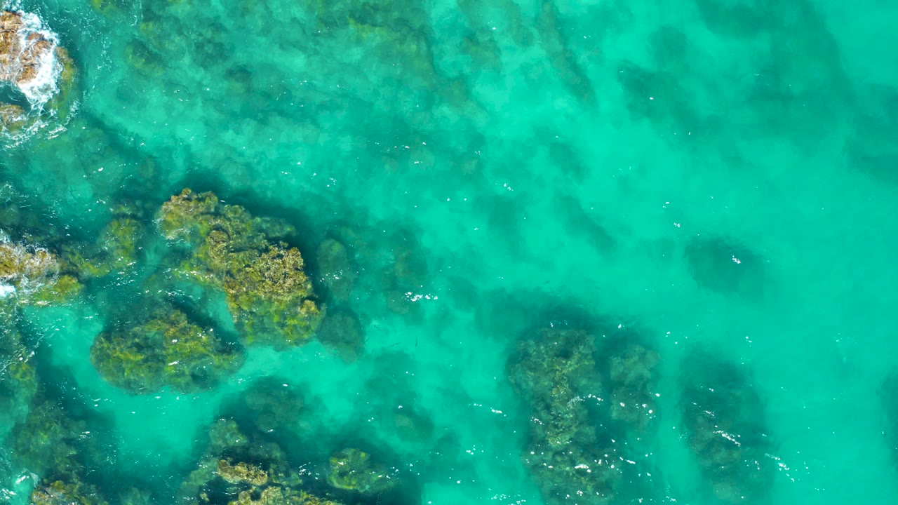 Overhead view in East Cape BCS Mexico showing turquoise water over dark rocks and green algae, with small white splashes at the upper left and no visible people.