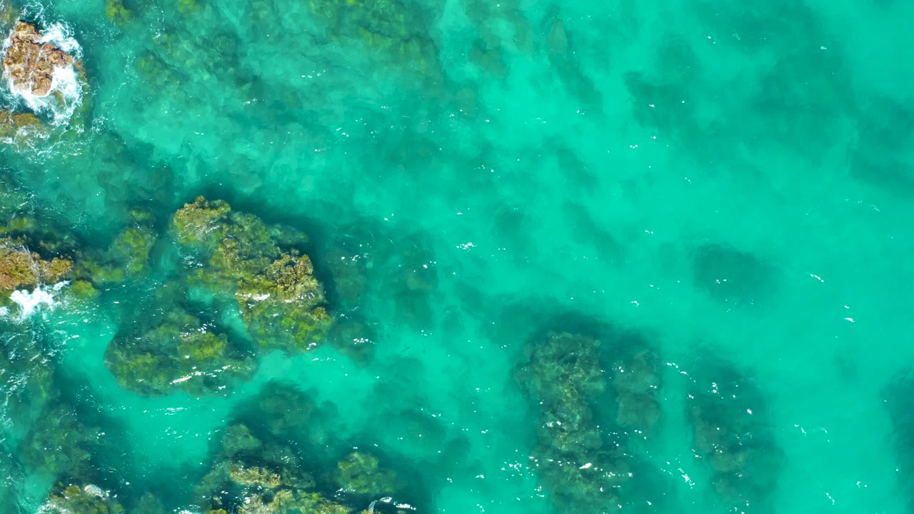 Aerial view in East Cape BCS Mexico with several dark submerged rocks visible beneath turquoise water, white streaks from wave motion, and no visible people.