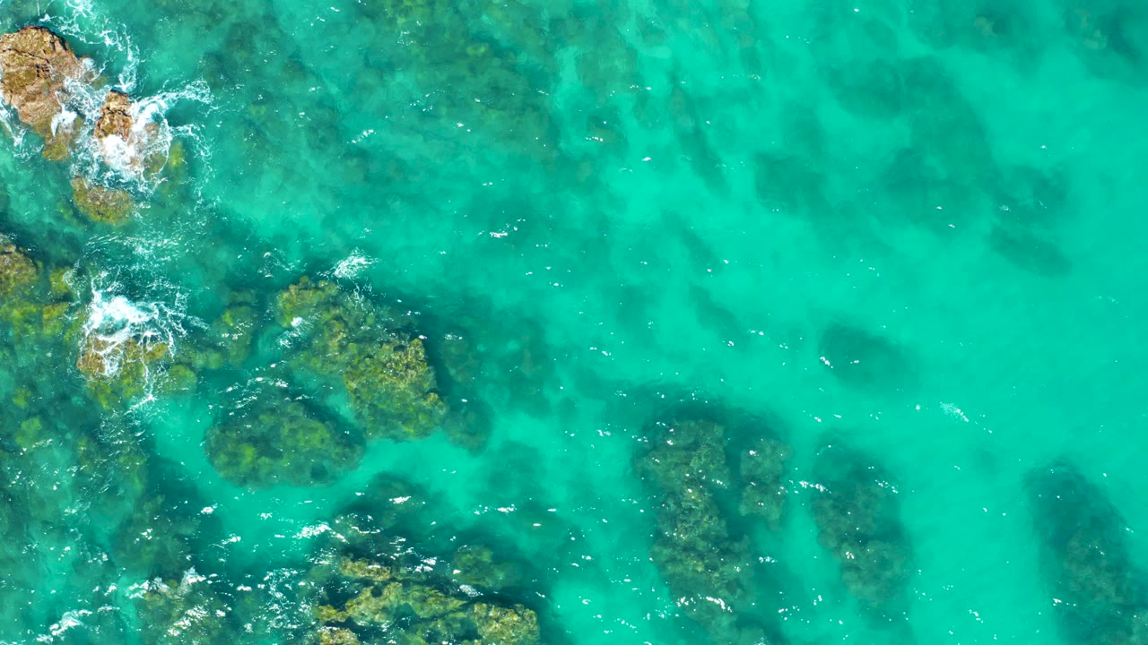 Overhead view of East Cape BCS Mexico turquoise shallow water, with submerged rocks along the left side and small white foam at the upper-left rocks, no visible people.