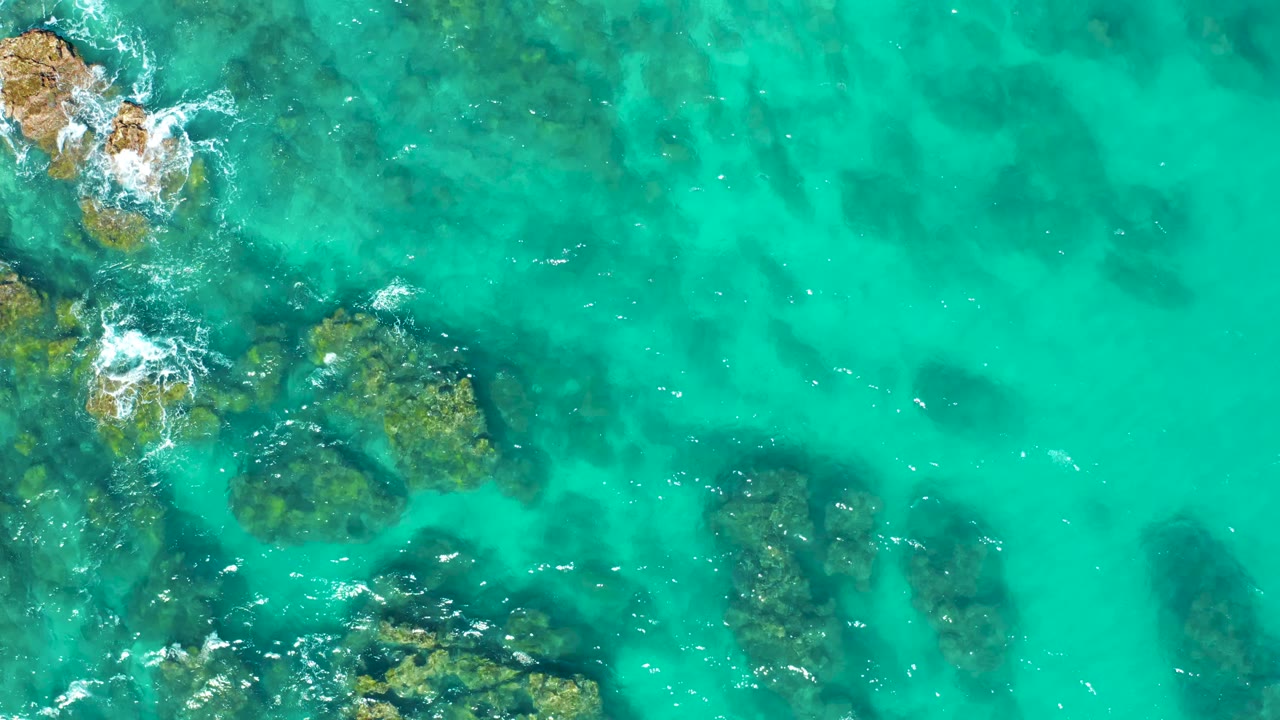 Aerial frame over East Cape BCS Mexico shallow turquoise water, where dark submerged rocks sit beneath the surface and white foam curls around the upper-left rock group, no visible people.
