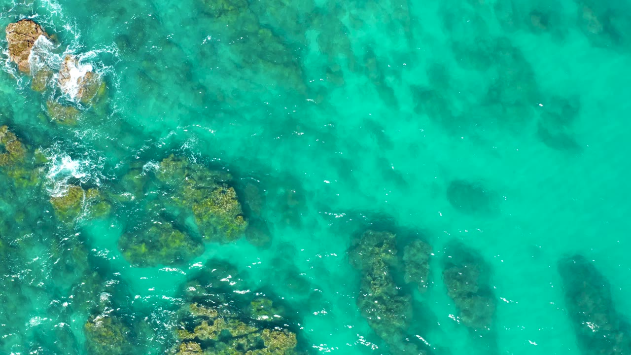 Overhead East Cape BCS Mexico water detail with blue-green shallows, submerged rock patches across the lower half, and brighter foam breaking along the left edge, no visible people.