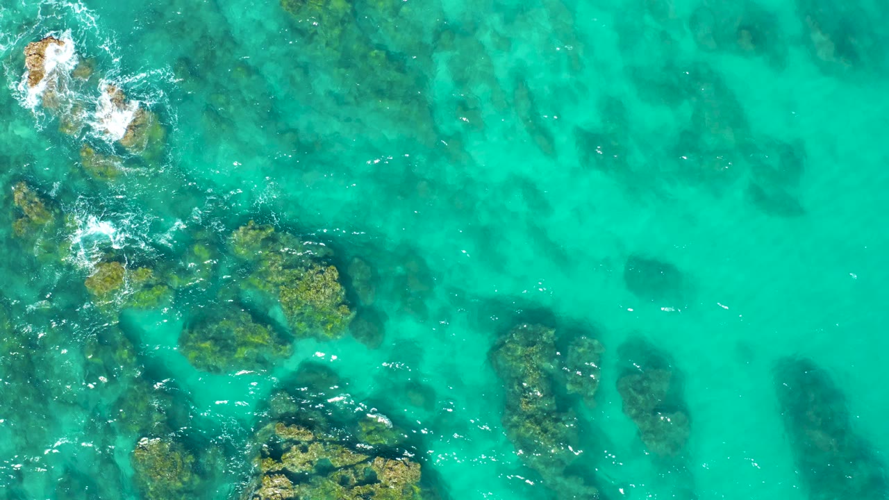 Aerial view in East Cape BCS Mexico with foamy exposed rocks near the upper left, darker seabed shapes under the turquoise water, and open clear water to the right, no visible people.