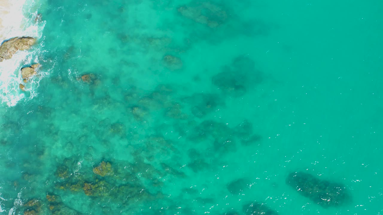 Aerial view of East Cape, Baja California Sur turquoise water with submerged rocks, a thin sandy shoreline at left, white surf near rocks, and no visible people.